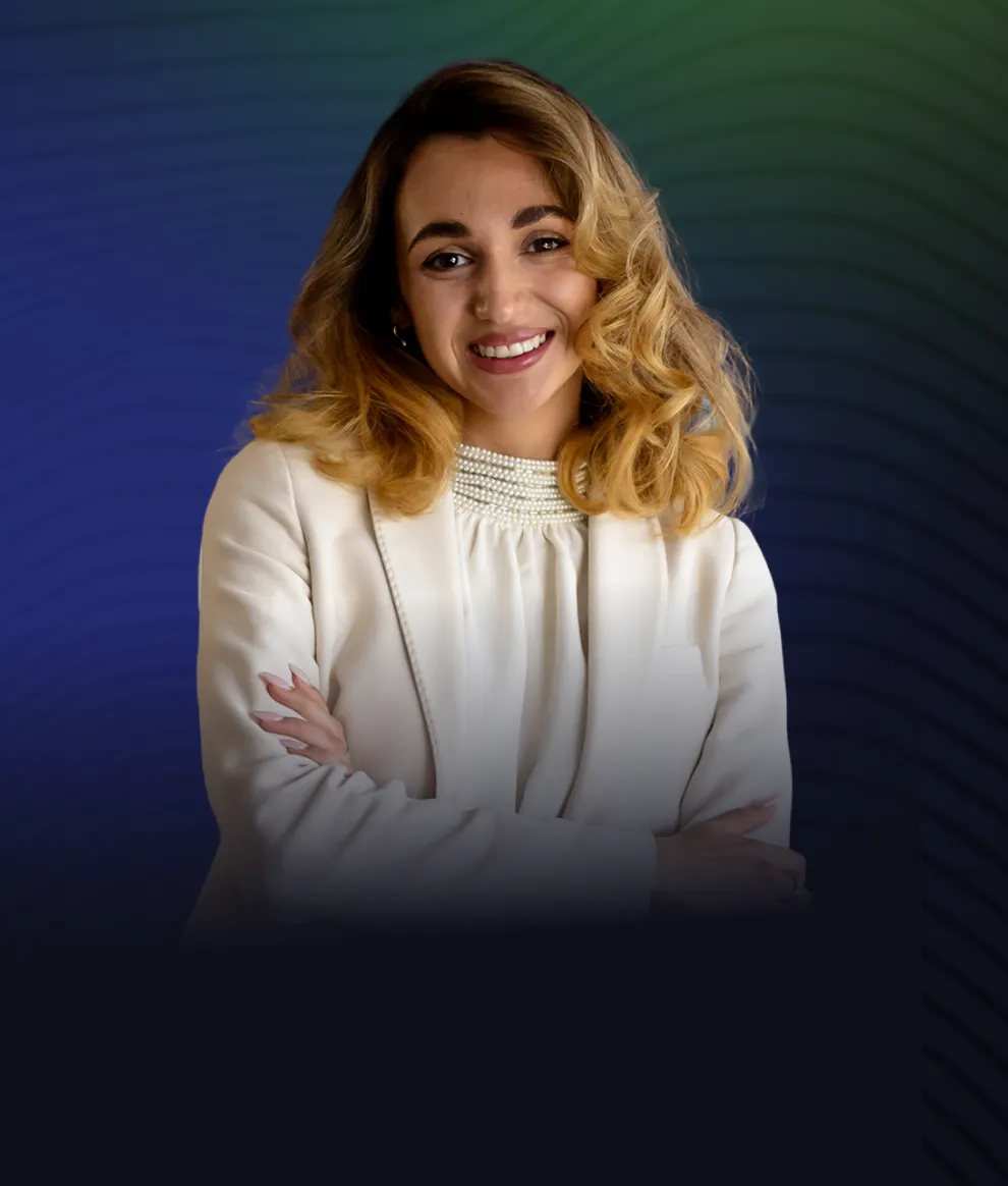 Smiling woman with blonde curly hair wearing a white blazer and pearl necklace, arms crossed.