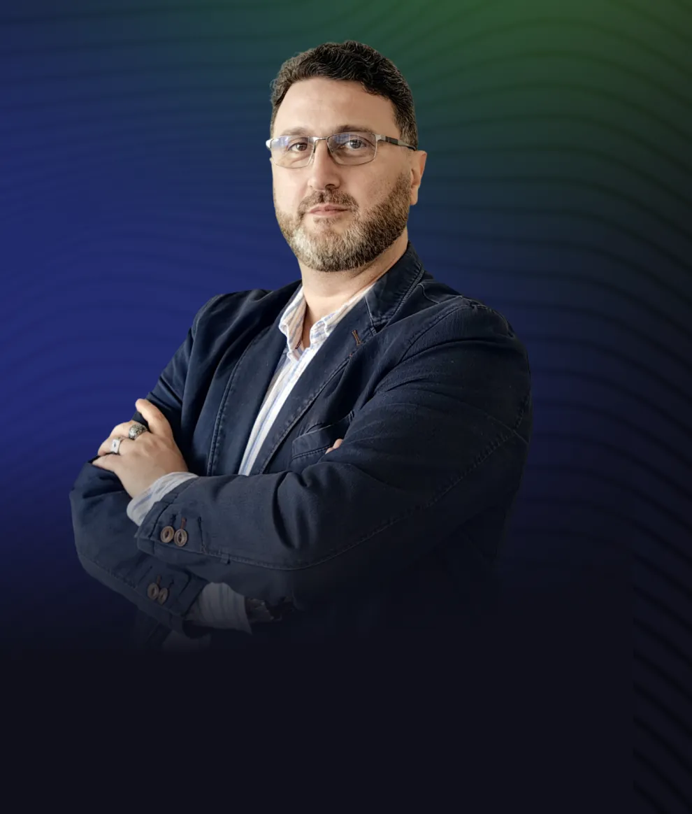 Man with glasses and beard wearing a dark blazer with arms crossed against a blue and green gradient background.