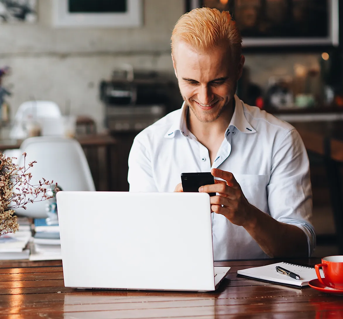 Smiling man in a white shirt using a smartphone while sitting behind a laptop at a wooden table with a notebook and red coffee cup.
