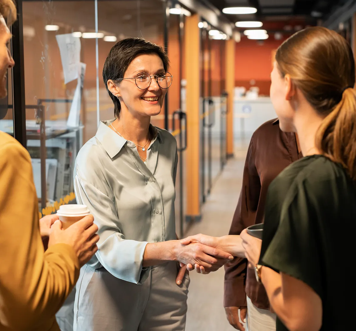 Two women shaking hands and smiling in a modern office hallway, with two other people nearby holding coffee cups.