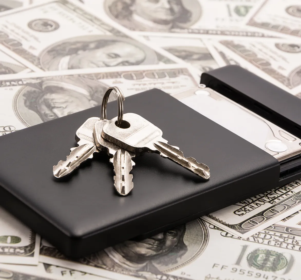 Set of three keys on a black external hard drive placed over scattered US hundred-dollar bills.