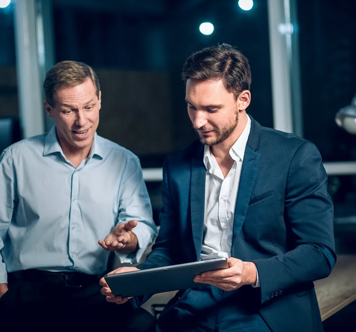 Two men in business attire discussing while looking at a tablet in an office setting at night.
