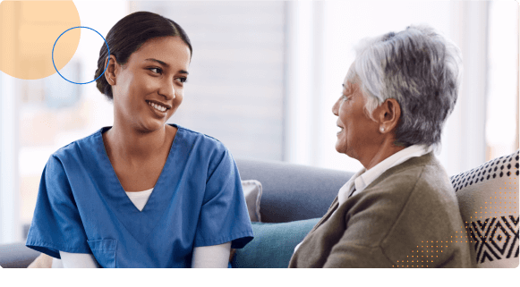 Smiling nurse in blue scrubs talking with elderly woman on a couch in a bright room.