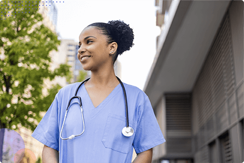 Smiling female nurse in blue scrubs with stethoscope standing outdoors near buildings and trees.