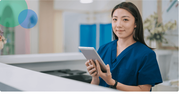 Smiling healthcare worker in blue scrubs holding a tablet at a reception desk.
