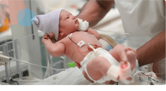 Newborn baby wearing a hat and connected to medical monitors, being held by a healthcare worker.