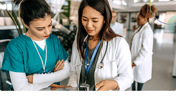 Two female medical professionals reviewing a tablet in a hospital corridor, another doctor walks in background.