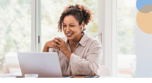 Smiling woman with earbuds using a laptop at a bright table in front of large windows.