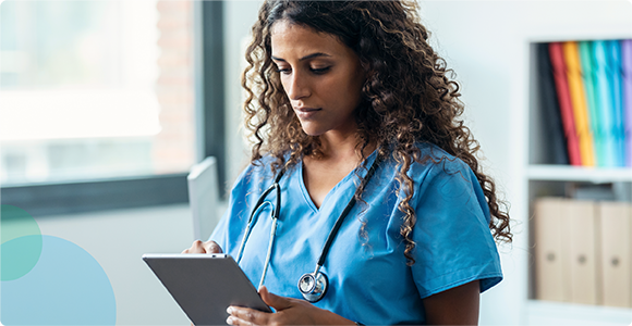 Female healthcare worker in blue scrubs reviewing notes on a clipboard in a bright office.