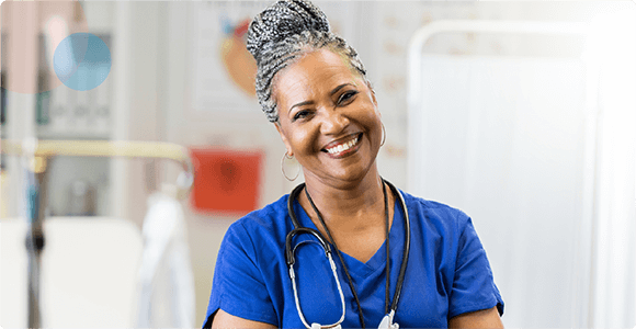 Smiling female nurse with gray braided hair wearing blue scrubs and a stethoscope in a medical setting.