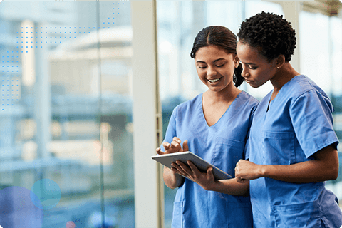 Two female healthcare workers in blue scrubs using a tablet by a large window.