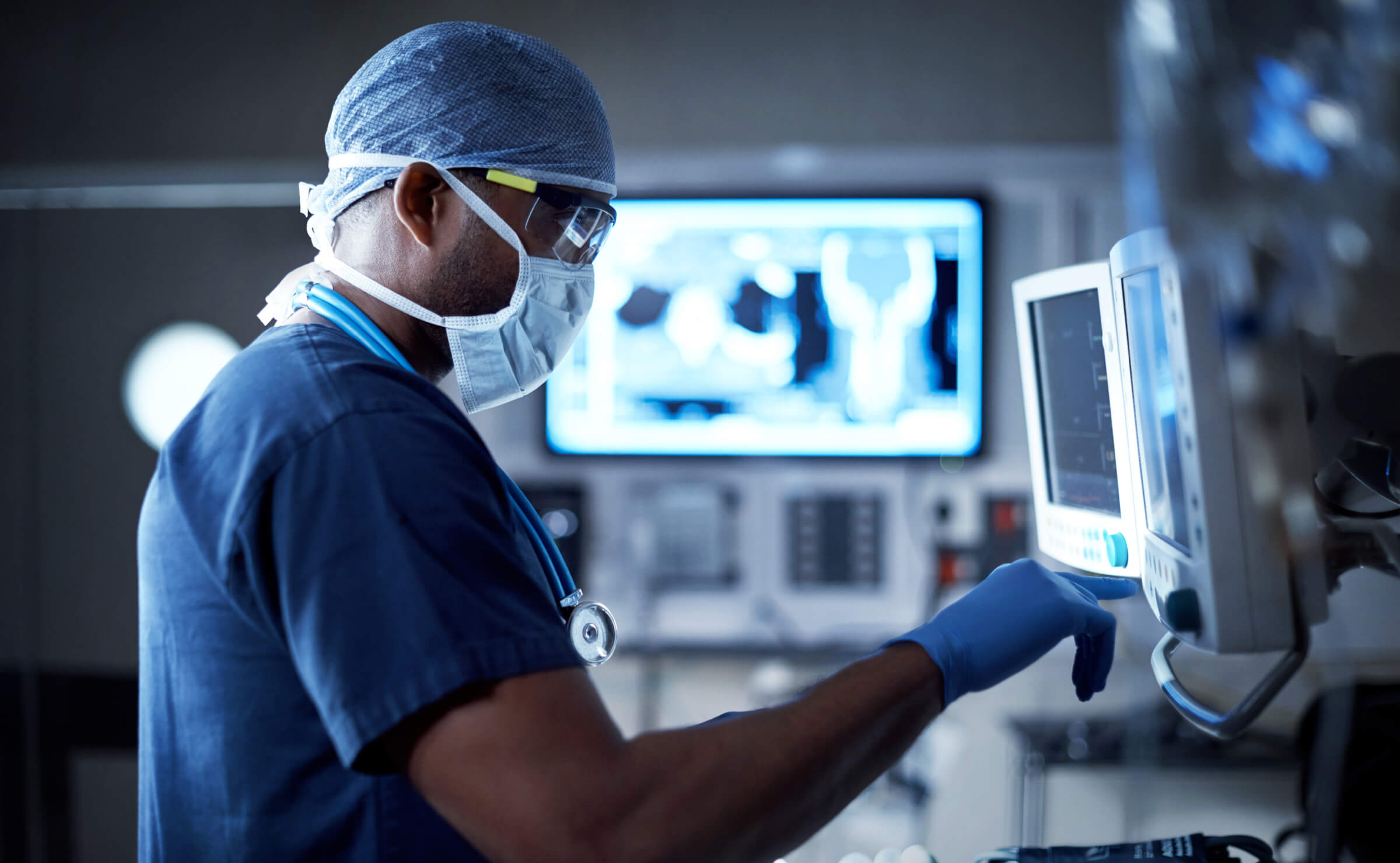 Masked doctor in scrubs and gloves adjusts a monitor in a dimly lit medical room.