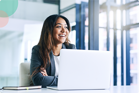 Smiling woman in a blazer sitting at a desk with a laptop in a bright modern office.