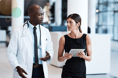 A male doctor in white coat talks with a woman holding a tablet in a modern office.