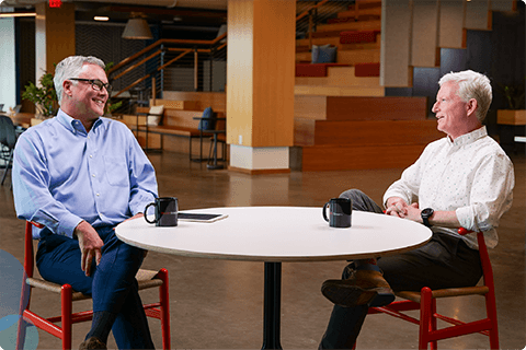 Two men sit at a round table in a modern office lounge, smiling and talking with coffee mugs.