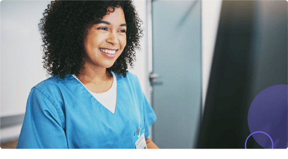 Smiling healthcare worker in blue scrubs looking at a computer screen in a clinical setting.