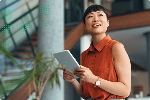 Woman in orange sleeveless blouse holds a tablet and looks upward, indoors by stairs with plants.