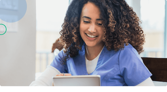 Smiling nurse in blue scrubs looking at a tablet in a bright room.