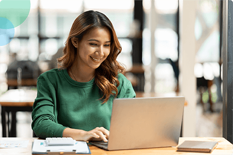 Smiling woman in green sweater working on a laptop at a table in a bright cafe.