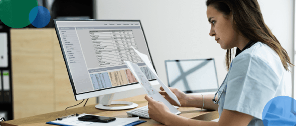 Medical professional reviewing documents while looking at a computer screen with data tables.