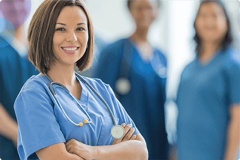 Smiling nurse in blue scrubs with stethoscope, other medical staff blurred in background.