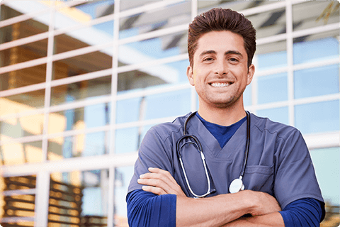 Smiling male doctor with stethoscope wearing scrubs standing with arms crossed outside a glass building.