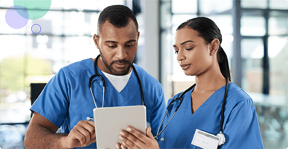 Two healthcare workers in blue scrubs reviewing information on a tablet in a hospital setting.