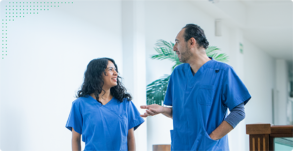 Two healthcare workers in blue scrubs having a conversation while walking indoors.