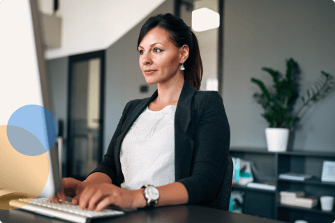 Professional woman typing on keyboard at desk in modern office with plants and shelves in background.