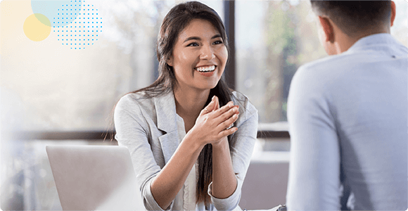 Smiling woman in gray blazer talking to a man in a bright office with a laptop nearby.
