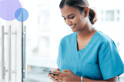 Smiling woman in blue medical scrubs using a smartphone indoors near a glass door.