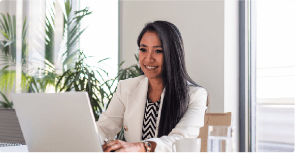 Smiling woman in white blazer working on a laptop at a bright office desk with plants behind her.