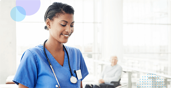 Smiling nurse in blue scrubs with stethoscope looking at a tablet in a bright medical facility.