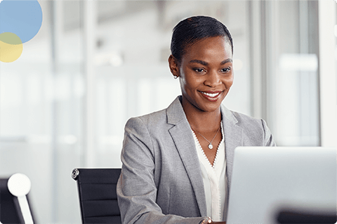 Smiling woman in gray blazer working on laptop in bright, modern office space.