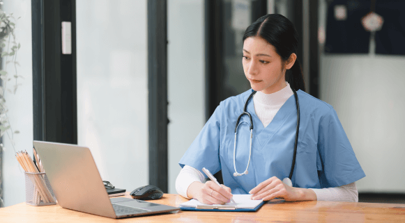 Female nurse in blue scrubs writing on a clipboard beside a laptop at a desk in a medical setting.