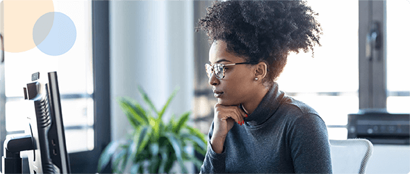 Woman with glasses concentrating on a computer screen in a bright office with a plant nearby.