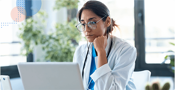 Female doctor wearing glasses and a stethoscope, focused on work at her laptop in a bright office.
