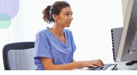 Healthcare worker in blue scrubs typing on a keyboard while looking at a computer monitor.