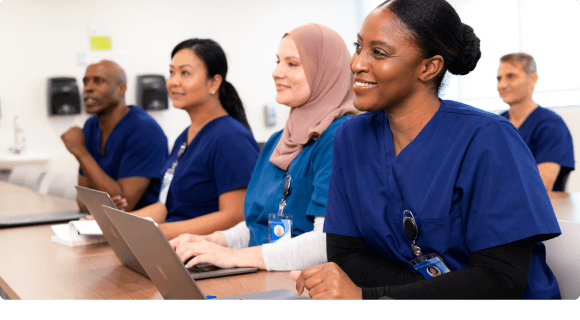 Smiling healthcare professionals in blue scrubs sitting at a table with laptops in a classroom setting.