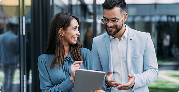 Smiling man and woman standing outdoors by glass building, looking at tablet in woman’s hands.