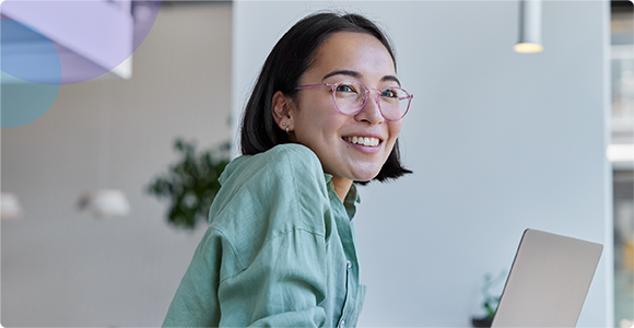 Smiling woman with glasses wearing a green shirt working on a laptop in a modern office.