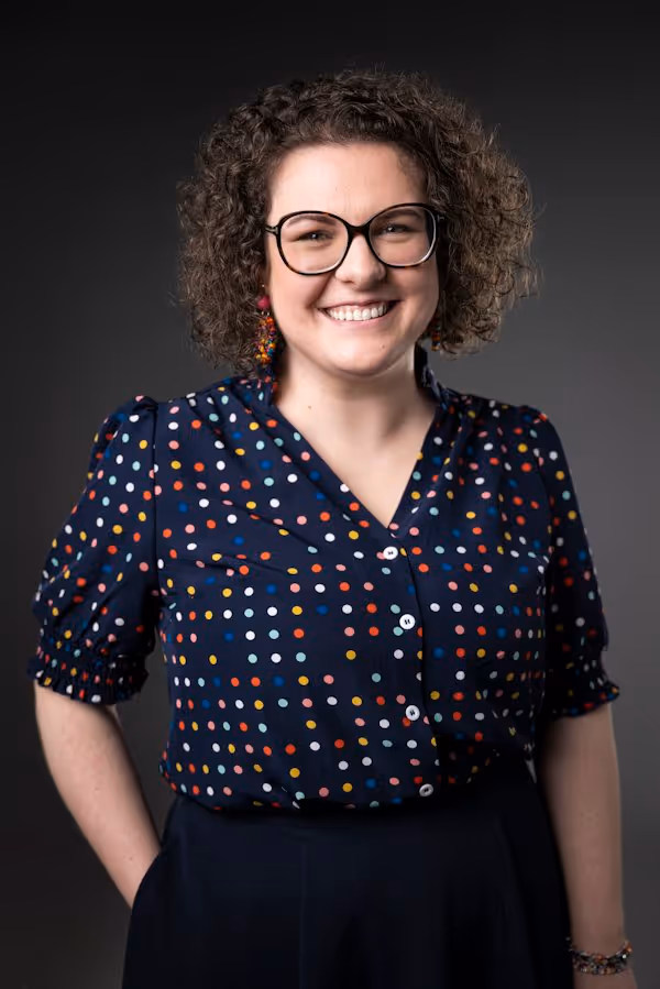 Porträt einer lächelnden Frau mit Brille und lockigen Haaren vor dunklem Hintergrund