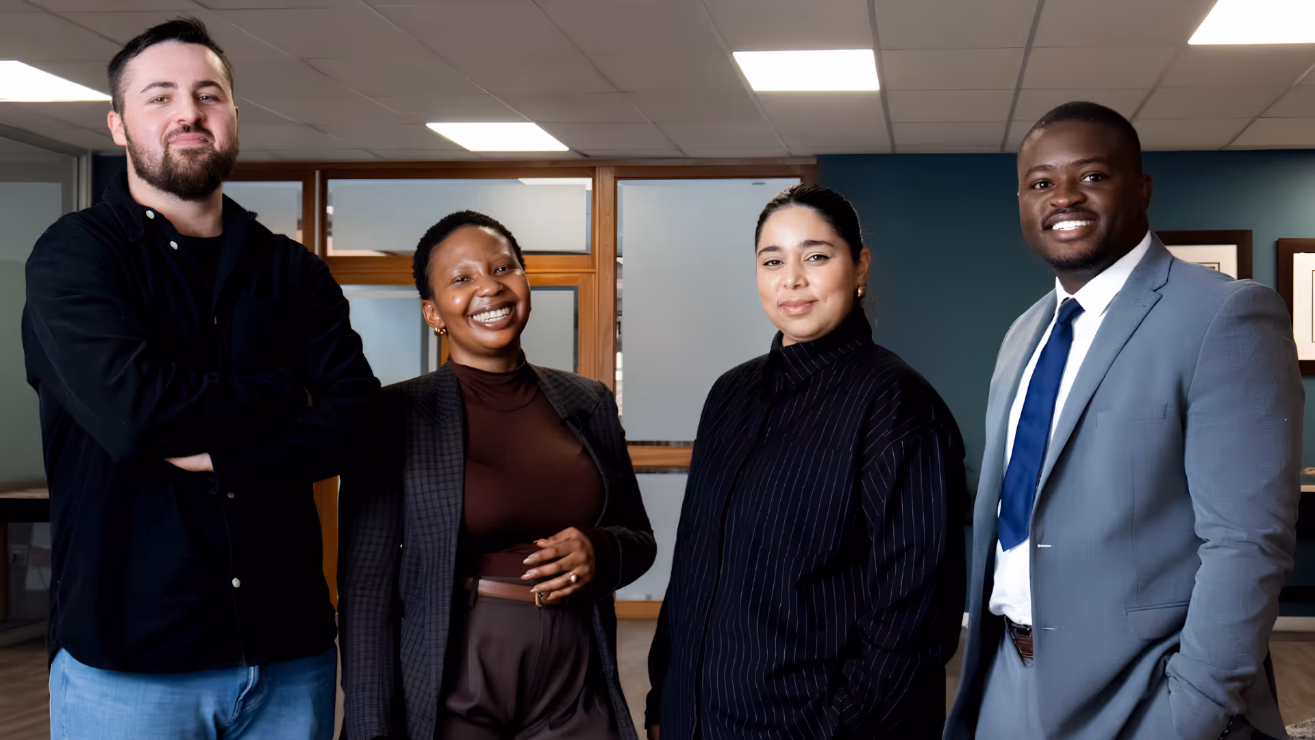 Four diverse professionals smiling and standing together in a modern office space.
