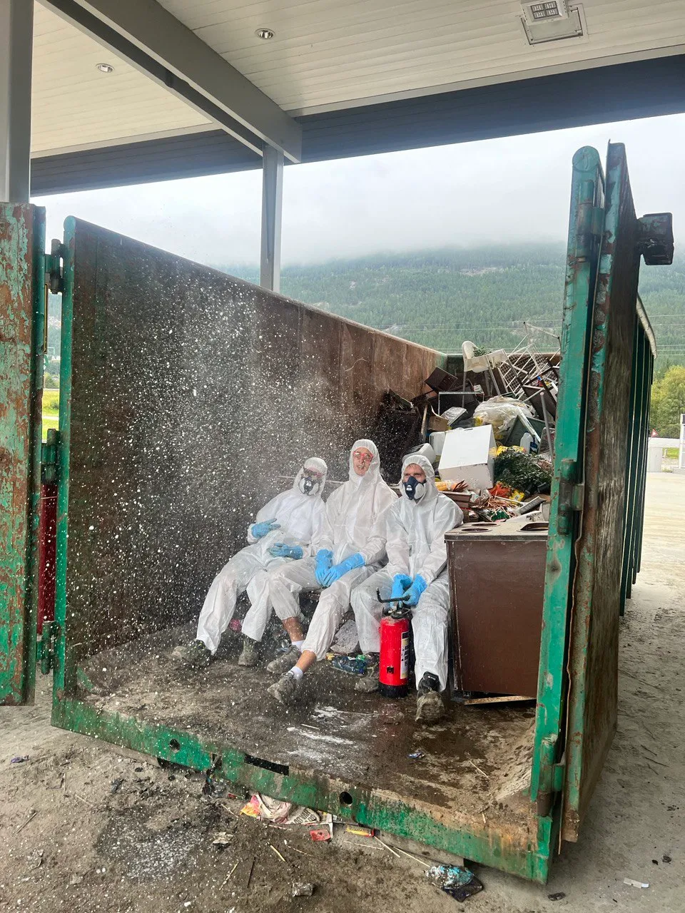 Tre personer i hvite vernedrakter sitter i en container med avfall
De blir sprayet med væske
