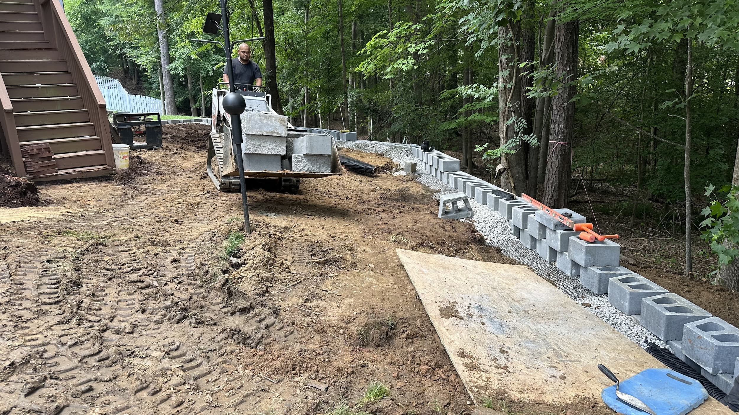 Man operating a small tracked vehicle carrying concrete blocks near a partially built retaining wall by a wooded area.