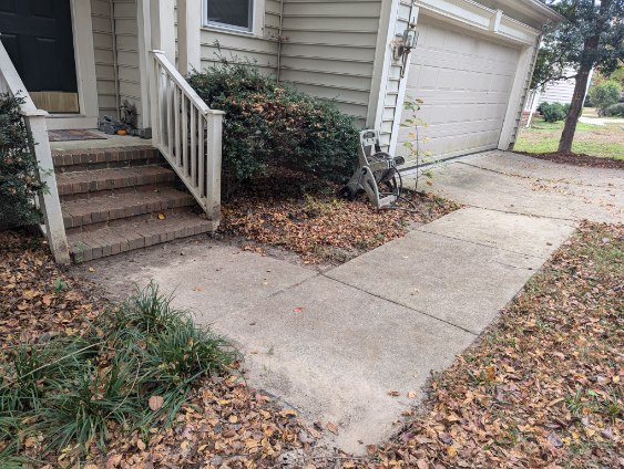 Sidewalk and driveway covered with fallen leaves outside a beige house with brick steps leading to a black door.