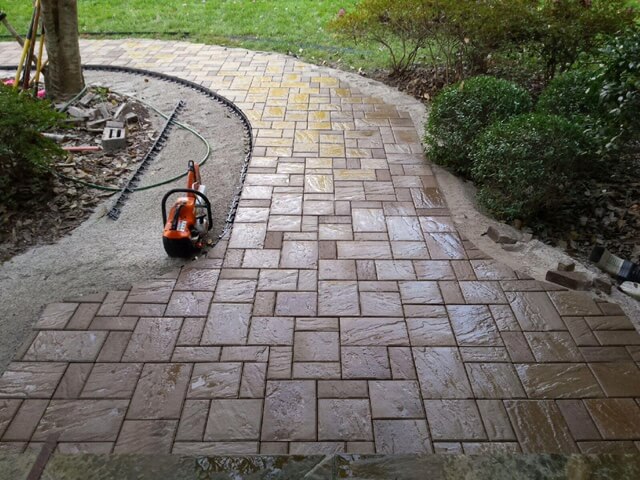 Partially installed curved brick walkway with wet pavers, edging materials, and a chainsaw placed on the ground.