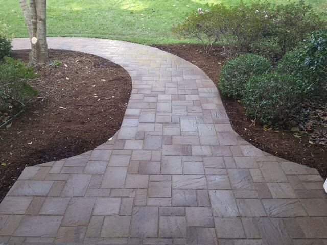 Curved stone paver pathway bordered by mulch, bushes, and grass in a garden.