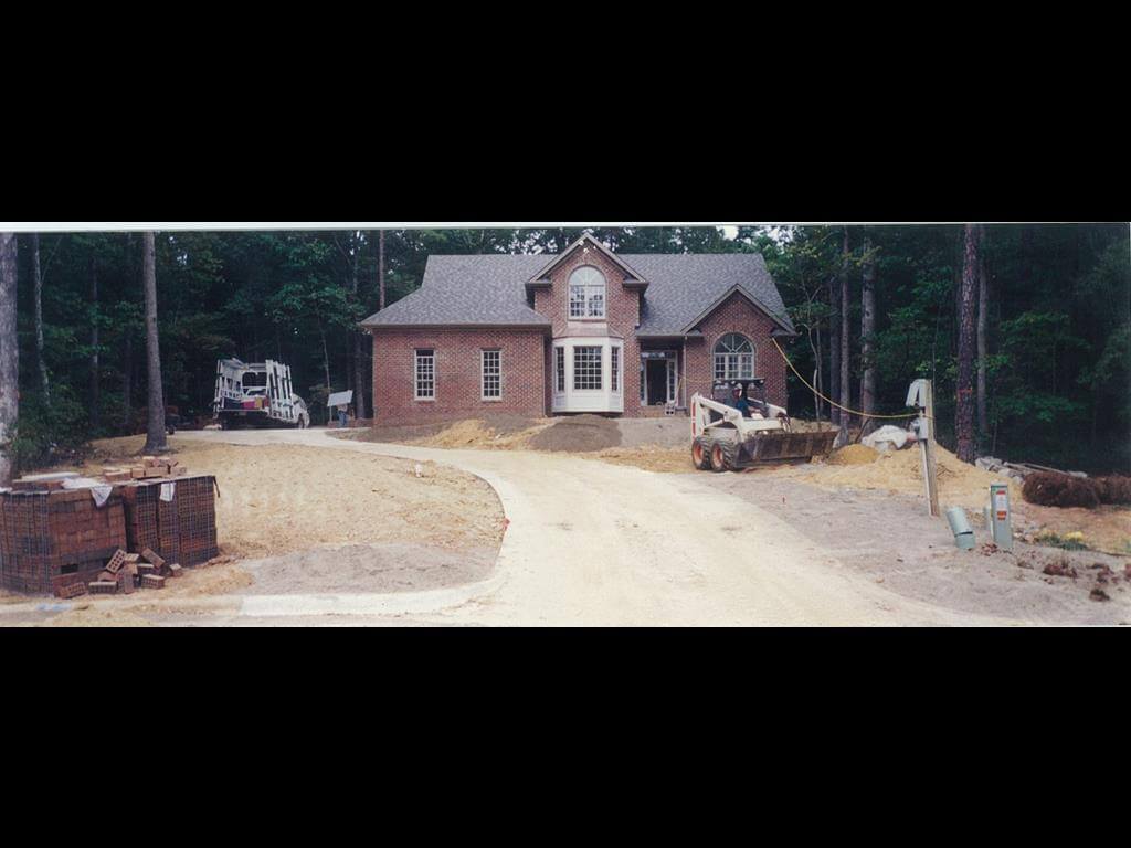 Brick house under construction with dirt driveway, a skid-steer loader working on landscaping, and stacks of bricks on the left side.