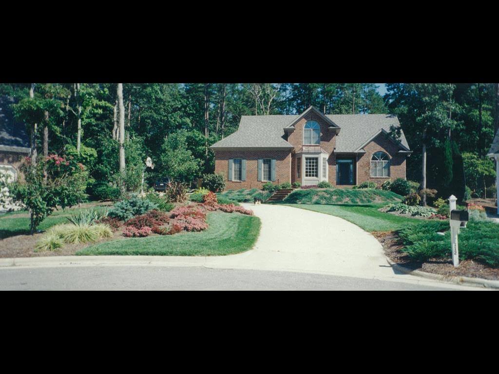 Large brick house with arched windows, a concrete driveway, manicured lawn, and well-maintained shrubbery and flower beds.